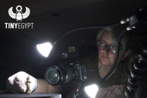 Julia Thorne photographing an Egyptian amulet. In the top left corner is the Tiny Egypt logo - a winged scarab holding a camera aperture, with the words 'tiny egypt' underneath