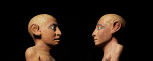 The head and shoulders of an ancient Egyptian wooden statuette of a man, shown twice in profile. The two images are facing each other, looking towards the centre of the image.